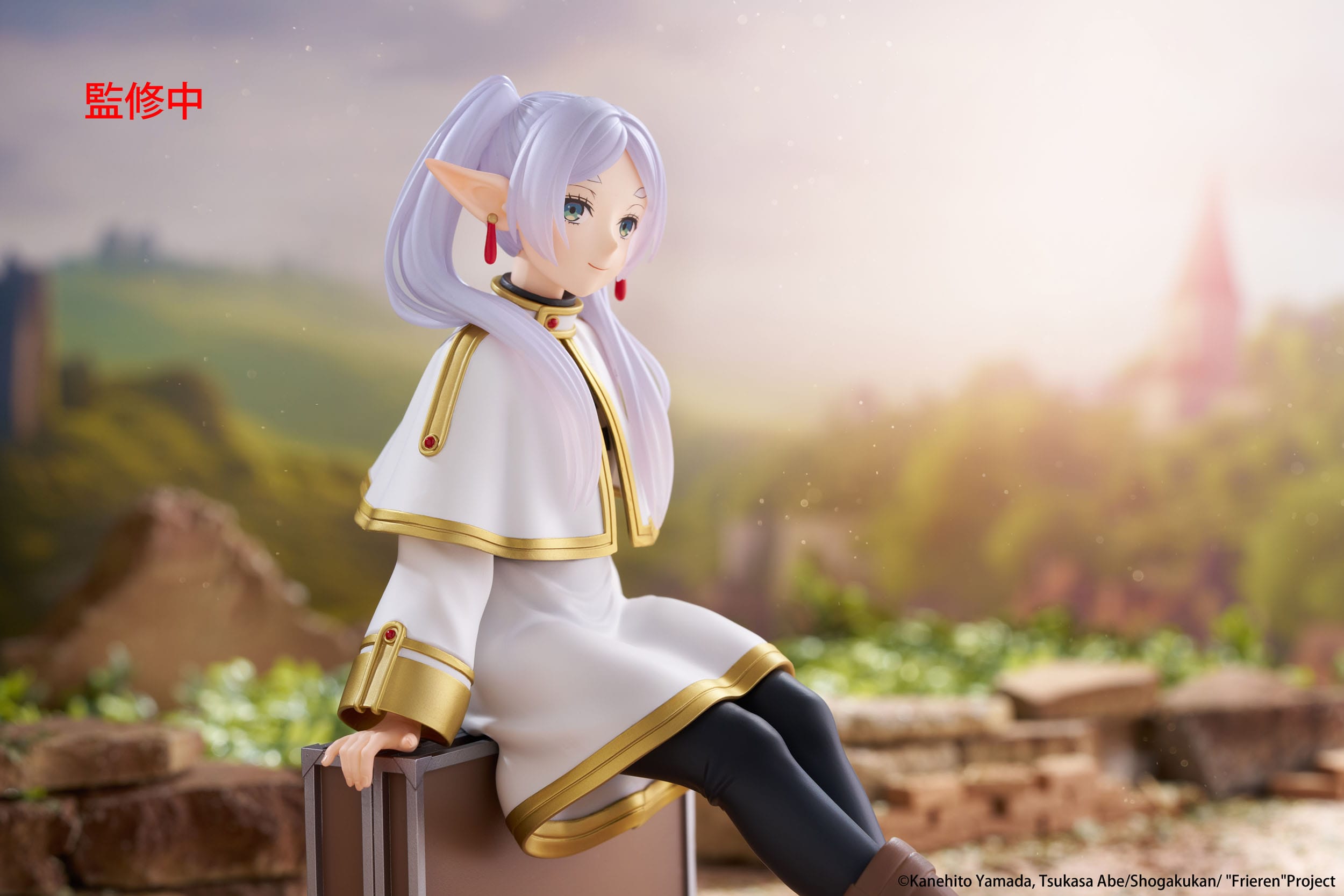 Anime character figurine with white hair and a white outfit with gold accents, sitting on a stone wall with a blurred scenic background.