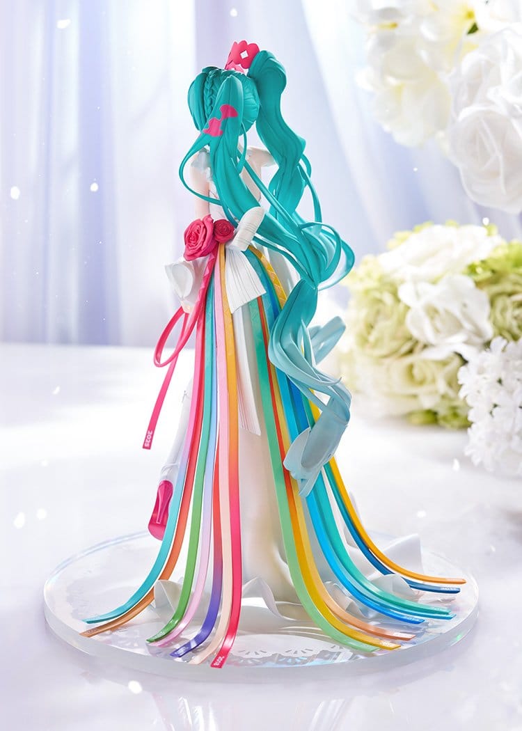 A decorative figurine or cake topper shaped like a woman with long teal hair and flowing multicolored ribbons, displayed on a clear round base against a soft white floral background.