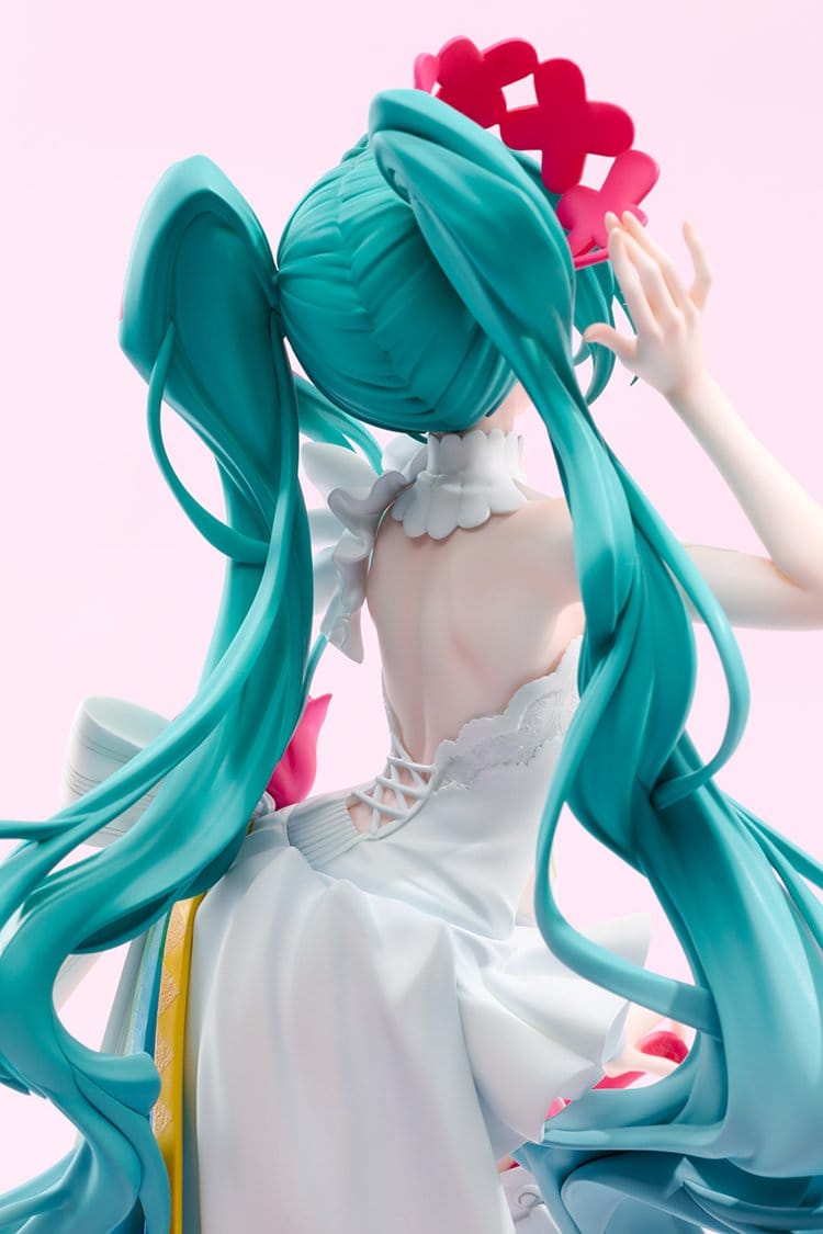 A close-up of a Hatsune Miku figure from behind, showcasing her signature teal twin-tails, white dress, and pink hair accessory against a solid light pink background.