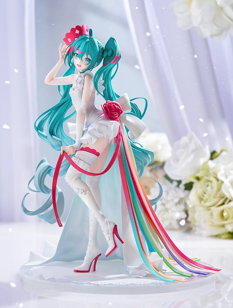 A collectible figurine of Hatsune Miku in a white wedding dress with long teal hair, pink flowers, and multicolored ribbons trailing from her dress, posed on a white base against a soft, blurred background with white roses.